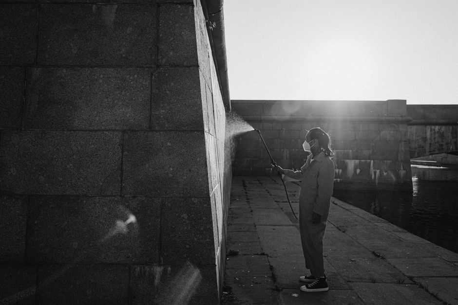 A person in a jumpsuit power washing a canal wall wearing a face mask, under bright daylight.